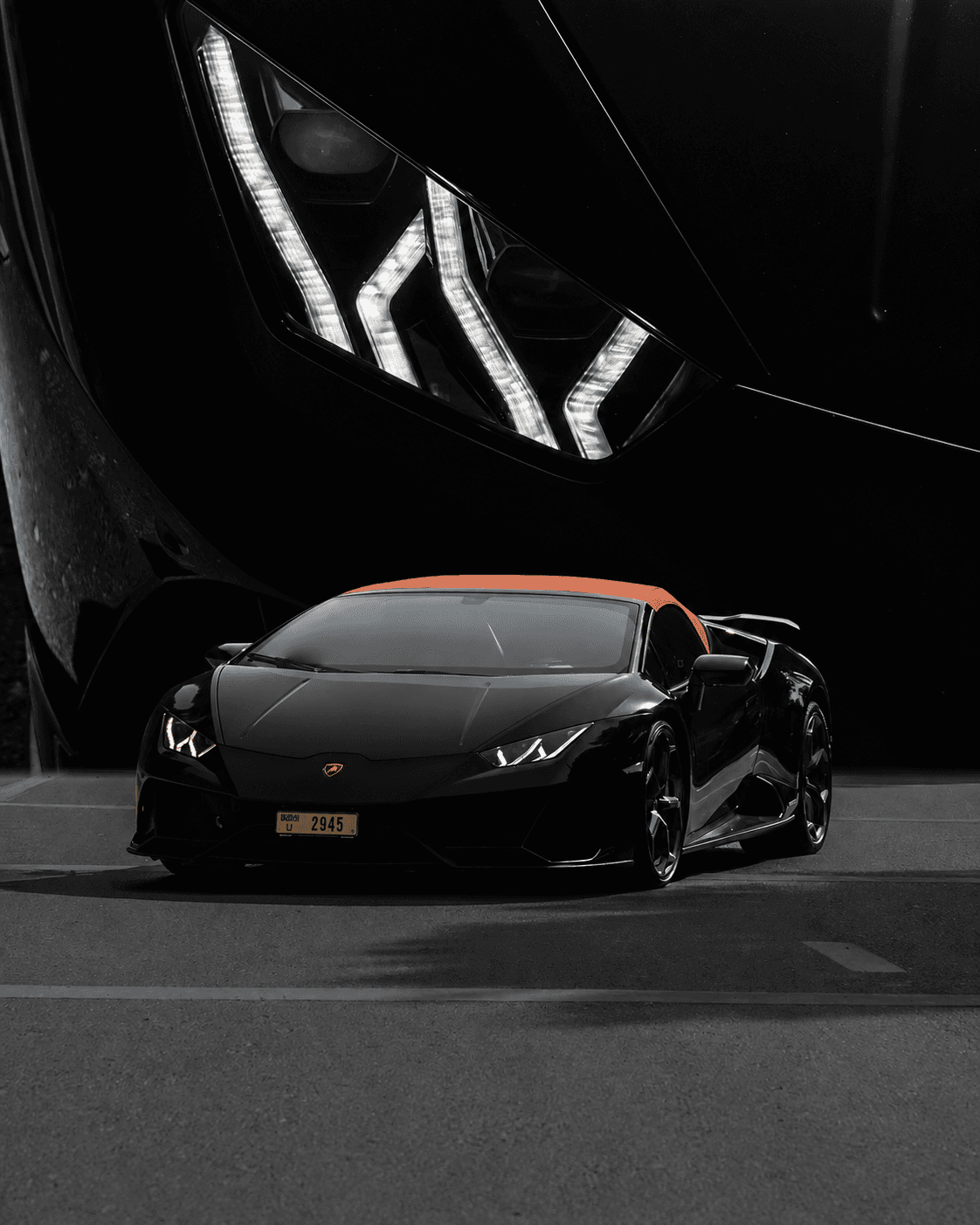 Black Lamborghini with an orange roof beneath a dramatic close-up of its headlight